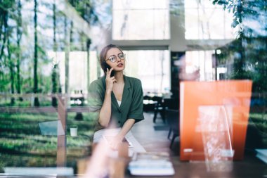 Modern salonda otururken gözlüklü, akıllı telefonla konuşan ve tablet tutan genç bayan öğrencinin cam manzarası.