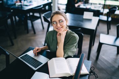 Yukarıdan, gözlüklü, masa başında oturan, dizüstü bilgisayarda çalışırken kameraya bakan ve ders kitabı okuyan neşeli genç bir kadın.