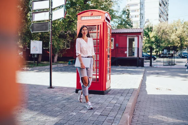 Full length portrait of positive hipster girl enjoying travel journey in England capital - London walking near red phone box with laptop computer, happy female tourist with netbook near telephone