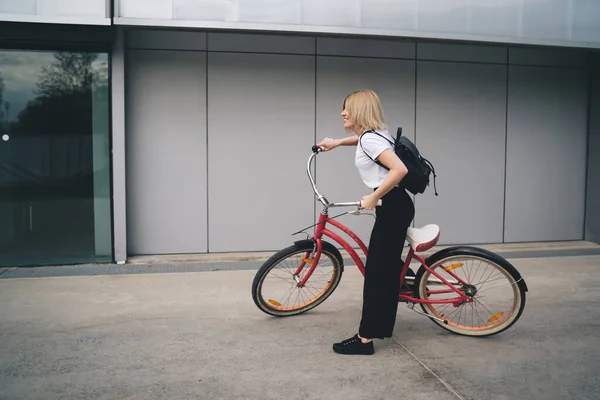 Side view of trendy blond woman with short hair carrying backpack while while riding pink bicycle against modern building exterior looking away