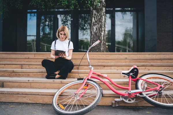 Casual young blond woman with black backpack sitting on wooden stairs on street with bicycle near and browsing modern tablet