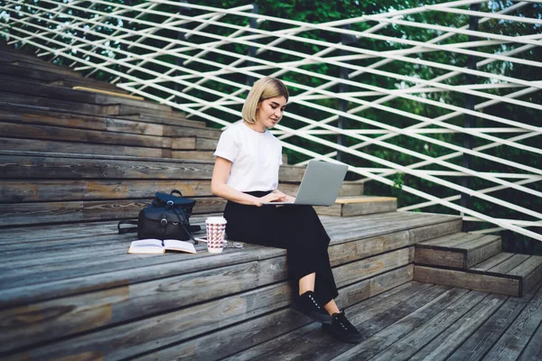女性の笑顔をバックパック付きの公式服で眺め 木製の階段に座っている間にノートパソコンに焦点を当て 対話するテイクアウトドリンク — ストック写真