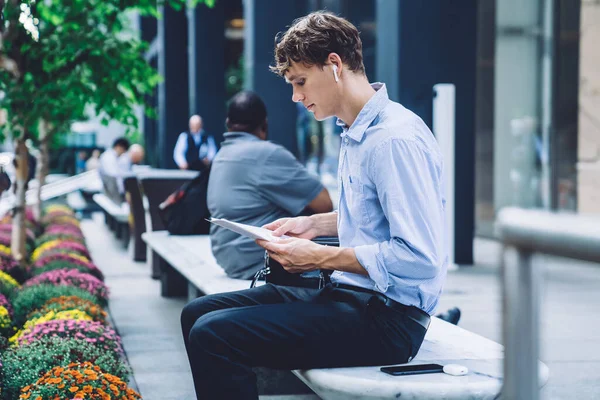 Clever male corporate economist doing accounting with papers sitting at urban setting in downtown and listening audio book via electronic earbuds, formally dressed man checking information in report