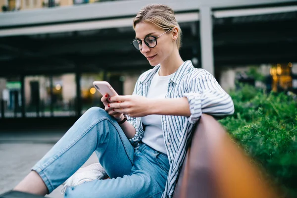現代的なスマートフォンを使用してラウンド眼鏡をかけカジュアルな服で魅力的なブロンドの女性の側面ビューとぼやけた通りの背景に緑の茂みの近くのベンチに座っている間笑顔 — ストック写真
