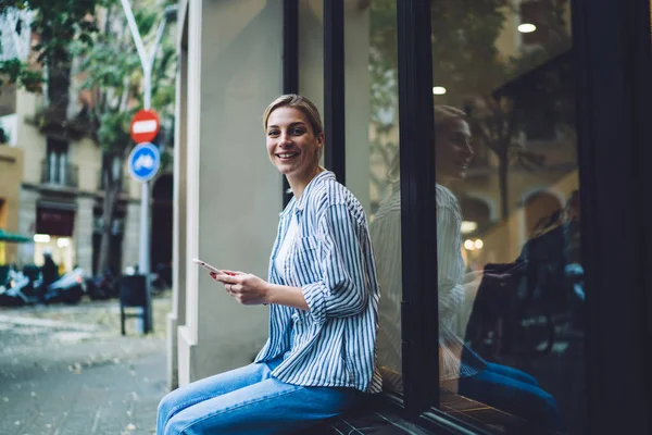 街の通りのカフェの外の窓の近くにスマートフォンに座ってメッセージング流行の服で幸せなブロンドの女性の側面図 — ストック写真
