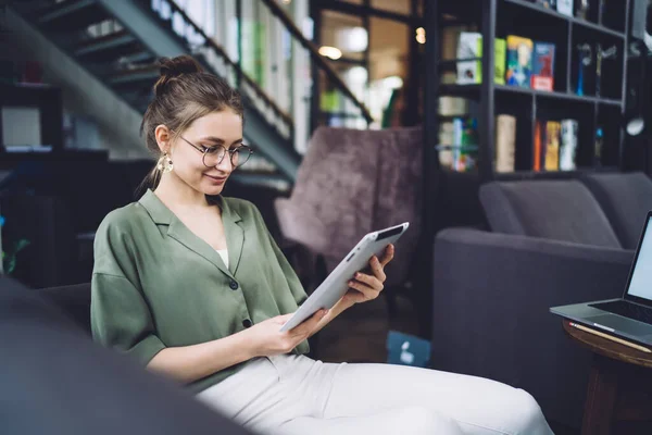 Young cheerful lady in casual wear and trendy glasses sitting at home and looking on tablet while watching video and smiling