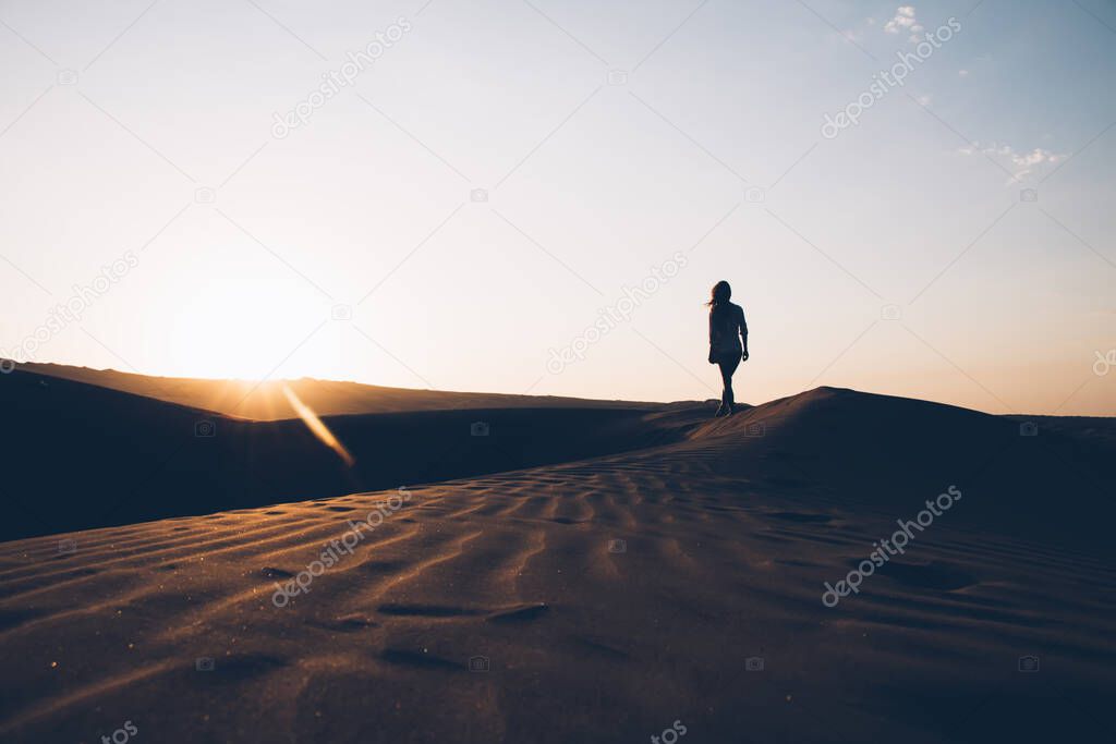 Calm peaceful Moroccan desert landscape with sun setting over horizon and illuminating rippled sand dunes and female silhouette against clear light sky 