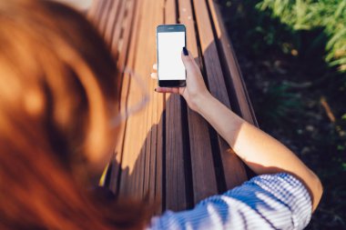 Kırpılmış, kızıl saçlı, sıradan giyinmiş, telefon kullanarak sosyal medyada arkadaşlarıyla sohbet eden, ahşap bankta oturan kadınların arka plan görüntüsü.