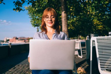 Neşeli kızıl saçlı kadın, görüş koruması için göz giysisi takıyor video izliyor ve laptopta haber okuyor güneşli bir günde dışarıda oturuyor, dijital göçebe bir kadın uzaktan çalışıyor.