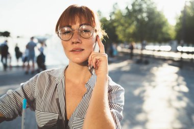 Şık çizgili tişörtlü ve gözlüklü çekici kızıl saçlı kadın şehir sokağıyla konuşurken kameraya bakıyor. 