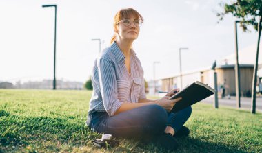 Hüzünlü kızıl saçlı, günlük giysili, gözlüklü kız öğrenci yeşil çimlerin üzerinde ayaklı haçla oturmuş proje üzerine ders kitaplarıyla çalışırken başka tarafa bakıyor.