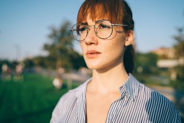 Ciddi kızıl saçlı, şık çizgili gömlekli, gözlüklü, bulanık arka planda yeşil parkı olan bir kadın. 