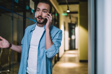 Gündelik giysiler içinde genç sakallı bir erkek akıllı telefondan konuşuyor ve ofis binasının koridorunda dikilirken haberlerle şaşırarak ellerini açıyor.