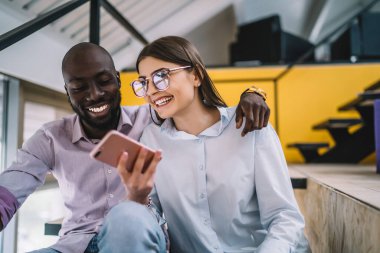 Neşeli genç kadın ve mutlu Afrikalı Amerikalı erkek cep telefonundan mesaj okuyor ve birlikte yaratıcı çalışma alanında otururken harika haberlerle gülüyorlar.