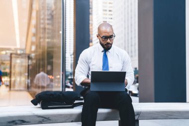 Gözlüklü dalgın Afrikalı Amerikalı yönetici taştan bir bankta oturmuş tabletle resmi kıyafetler giyiyor ve sosyal ağda mesajlaşıyor.