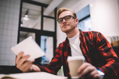Gündelik giysiler ve gözlüklerle yakışıklı genç adam kahve dükkanında kameraya bakarak bir kupa taze içecek ve telefon alıyor.