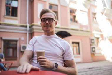 Tatmin olmuş beyaz gömlekli ve gözlüklü genç adam güneşli bir günde kafede dinlenirken bir fincan kahve içiyor.