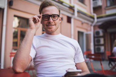 Neşeli, karizmatik genç adam arkadaşlarıyla sohbet ediyor. Boş vakitlerini telefon takıp, bardakları ayarlıyor ve yaz kafesinde kameraya bakıyor.