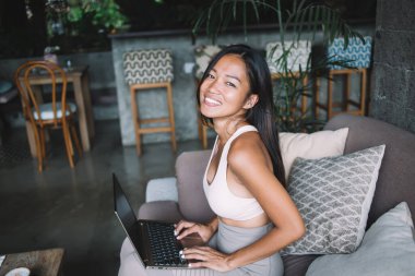 Spor giyim kuşamında, dizüstü bilgisayar kullanıp rahat bir kafede kanepede otururken kameraya bakan neşeli, genç ve çekici bir kadının yan görüntüsü.