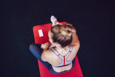 Yukarıdan gelen eşofmanlı genç bayan antrenman sırasında dinleniyor pilates stüdyosundaki telefonun yanında oturuyor.
