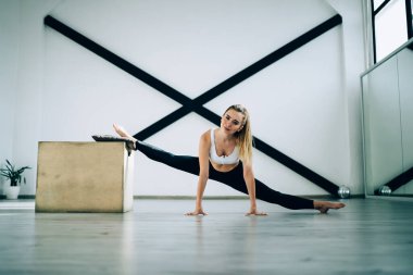 Spor kıyafetleri içinde, parlak, kararlı bir kadın kameraya gülümserken ön bölme egzersizi yapıyor ahşap kutuya ve yere yaslanıyor ve modern spor salonundaki siyah spor duvarına dayanıklı bantlara yaslanıyor.