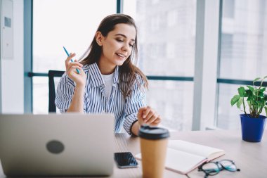 Modern ofisin penceresinde masa başında otururken günlük kıyafetlerle gülümseyen ve açık planlamacıda notlar okuyan mutlu genç kadın.