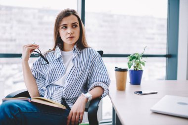 Şık giyinen düşünceli genç bayan öğrenci modern sınıfta kitap okurken sandalyeye oturup gözlerini kaçırıyor. 