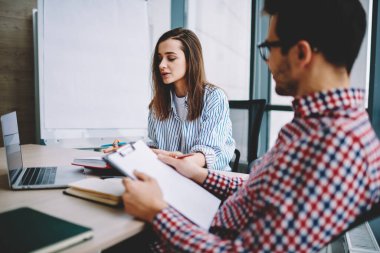 Erkek ve kadın iş ortakları gündelik giysiler içinde masa başında otururken ve modern ofiste belgeleri okurken iş teklifinde birlikte çalışıyorlar. 