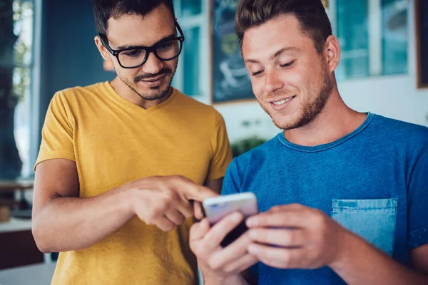 Joyful young man in casual clothes relaxing with content handsome friend in glasses pointing at screen of smartphone in cafeteria