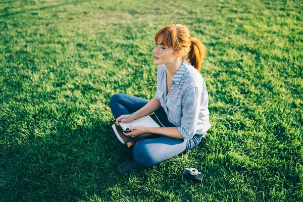 公園でノートを学びながら緑の芝生の上に足を組んで座ってカジュアルな服や眼鏡の赤い毛の小学生の女の子の上から — ストック写真