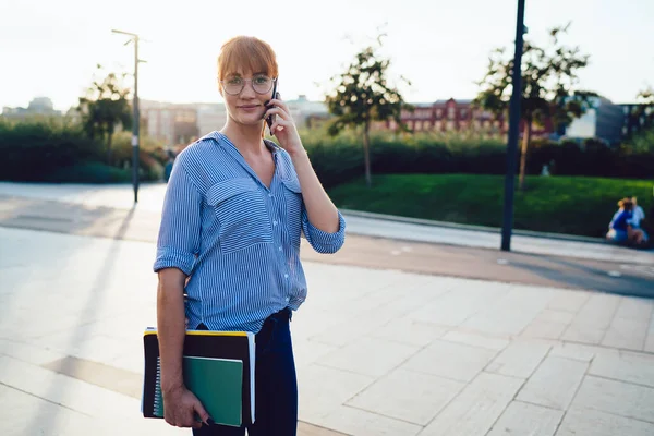 Half length portrait of good looking casual dressed hipster girl talking on mobile phone in roaming, attractive caucasian woman having smartphone conversation standing on street satisfied with tariffs