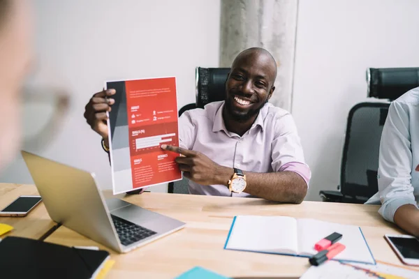 Cheerful African American man in elegant clothes pointing finger at document and explaining information to business partners while sitting at wooden table with papers and devices during conference in light modern office