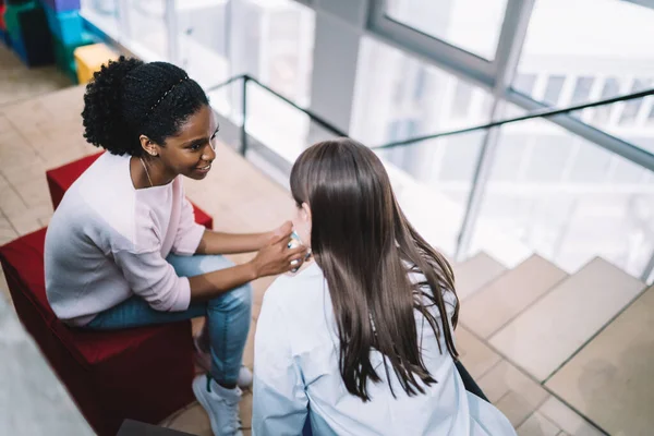 From above satisfied young diverse women in casual clothes talking and sharing news while sitting together on poufs near stairs in college