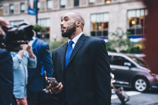 Successful bald and bearded African American entrepreneur in formal clothes standing and looking away while using smartphone outside on blurred background