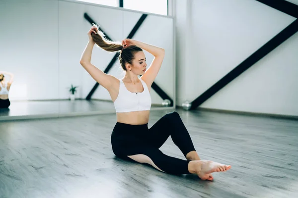 床に座って 大きな鏡でフィットネスジムで練習する前に髪を配置スポーツ衣装の若いスポーツ女性 — ストック写真