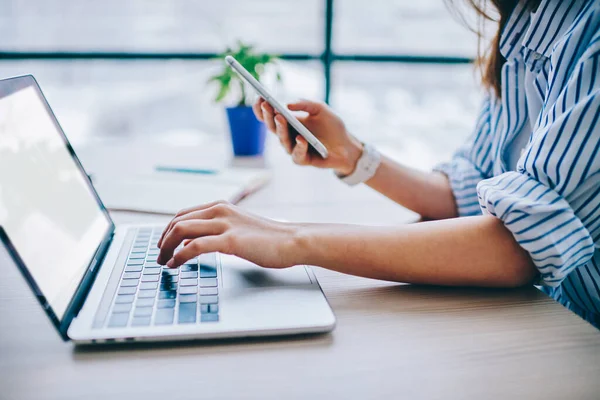 Crop hands of faceless female office employee in casual clothing typing on keyboard of empty screen laptop and surfing on mobile phone at modern office