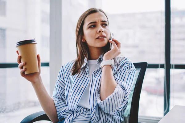 Serious female entrepreneur in stylish casual clothing drinking coffee to go while talking on mobile phone and looking away at modern office