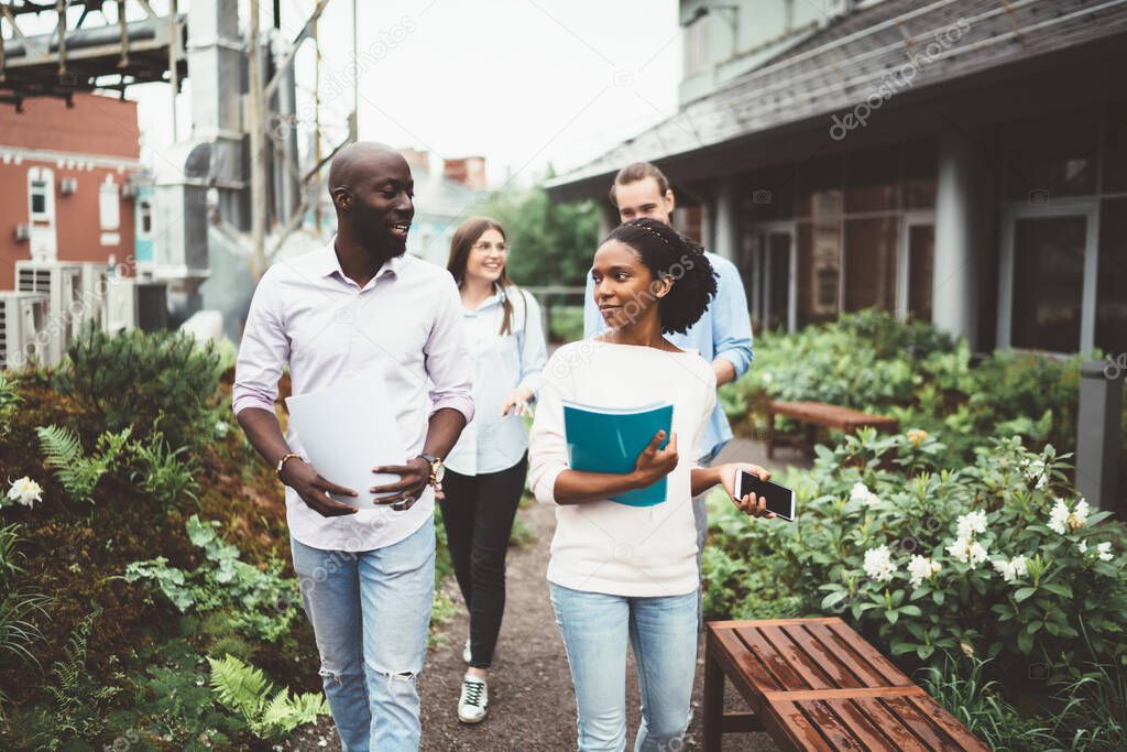 Content casual employees walking on terrace of modern building and chatting while carrying papers on background of cityscape