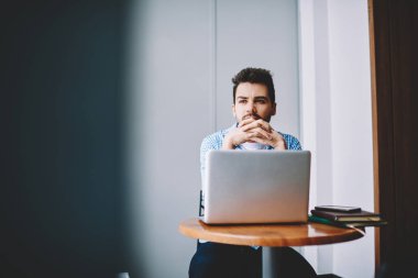 Ciddi odaklı, elleri bağlı genç bir adam, gündelik giysiler içinde modern dizüstü bilgisayarla çalışma masasında oturuyor ve gözlerini başka yöne çeviriyor.