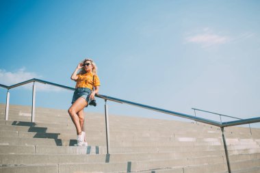 Tatilde, şık sarı saçlı, şık giyimli, güneş gözlüklü, trabzana yaslanmış ve arka planda mavi gökyüzüne bakan bir kadın. 