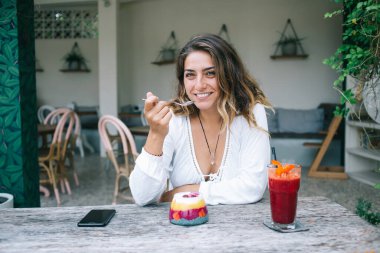 Aktif, kendine güvenen, dut gibi sulu jöle yiyen, kırmızı kokteylli masada oturan ve bahçedeki kafenin dışındaki kameraya bakan bir kadın. 