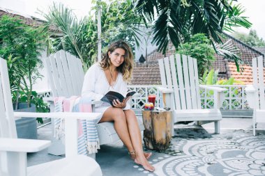 Çıplak ayaklı, beyaz elbiseli çekici bir kadın kameraya bakıyor ve gülümsüyor. Kırmızı meşrubatlı, renkli bir tatlı ve bulanık tropikal arka planda bahçede kitap okurken beyaz koltuğun yanında oturuyor.