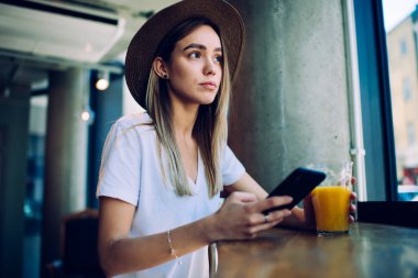 Kafe masasında oturmuş taze meyve suyunun tadını çıkarırken şapkalı, akıllı telefonlu ve bakışlarını kaçıran modaya uygun genç bir kadın.