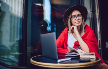 Şapkalı ve gözlüklü, renkli kırmızı battaniyeli bir kadın sokak kafesinde otururken kitap okuyor ve dizüstü bilgisayarda sörf yapıyor.