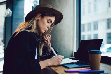 Çalışkan, stil düşkünü öğrencinin iş sırasında not defterine düşünceli bir şekilde yazıp kafenin yanındaki masada oturmasının yan görüntüsü. 