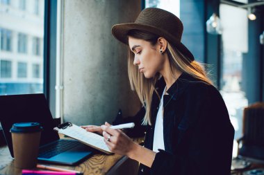 Kafe masasında dizüstü bilgisayarı ve kahveyle otururken panoda notları kontrol eden modaya uygun şapkalı ciddi genç bir kadının yan görüntüsü.