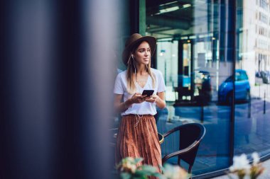 Şapkalı ve kulaklıklı düşünceli bir kadın. Sıradan giysiler içinde. Sokakta duruyor.