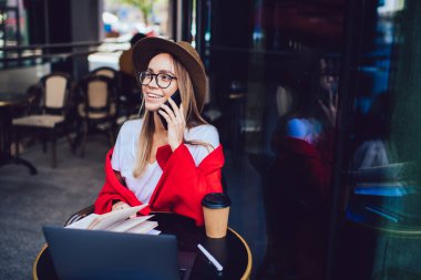 Moda şapkalı, gözlüklü, renkli kırmızı battaniyeli, akıllı telefonlu, dizüstü bilgisayarı ve kahve fincanı olan hoş bir kadın.
