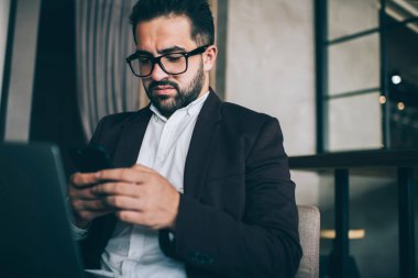 Gözlüklü ve siyah takım elbiseli yetişkin sakallı iş adamı akıllı telefon kullanıyor ve koltukta oturup dizüstü bilgisayarla çalışırken mesaj okuyor.