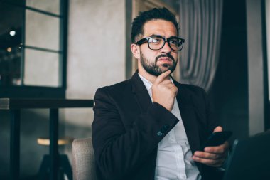 Resmi takım elbiseli ve gözlüklü düşünceli bir adam. Ortağıyla akıllı telefon mesajlaşması yapıyor. Çenesine dokunuyor.